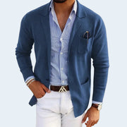 Man wearing a blue blazer over a striped shirt with white pants on a light gray background