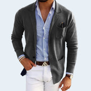 Man wearing a gray blazer over a blue striped shirt with white pants on a light gray background