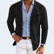 Man wearing a black blazer over a blue striped shirt with white pants on a light gray background