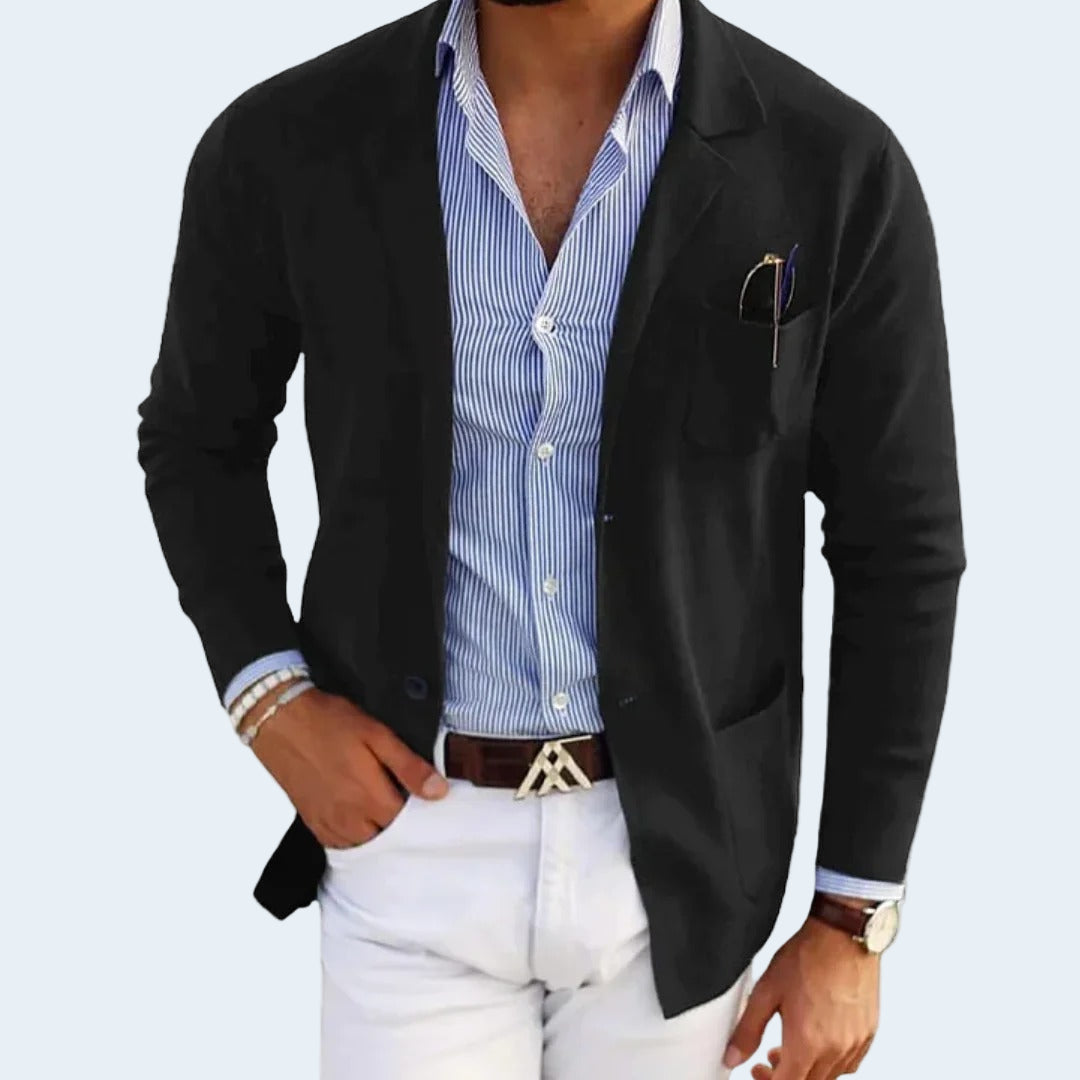 Man wearing a black blazer over a blue striped shirt with white pants on a light gray background